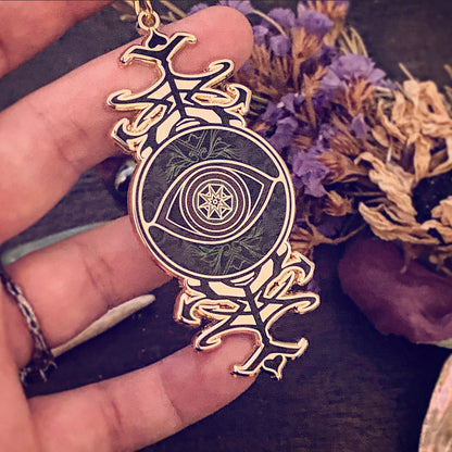 A gold-toned pendant featuring a central eye design with a star and crescent moon motif, held in a hand against a dark background with dried flowers.