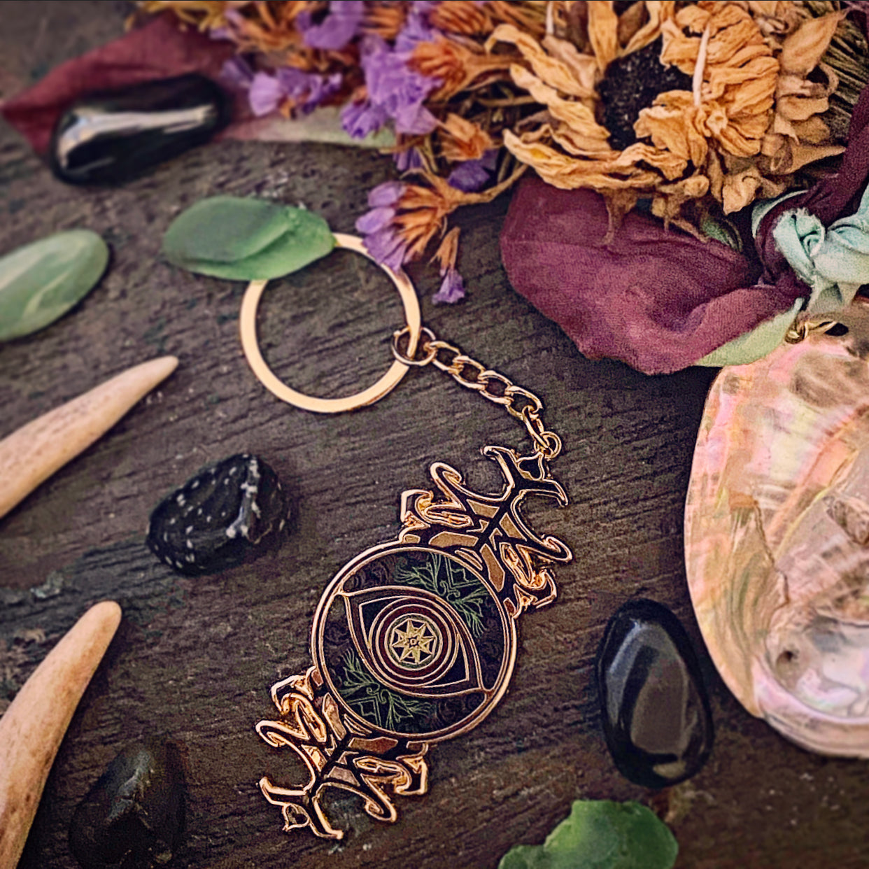 A decorative keychain with a circular metal ring and chain, featuring an ornate gold-toned pendant with a central eye motif and green botanical details, styled on a dark wooden surface with dried flowers, stones, and a shell.