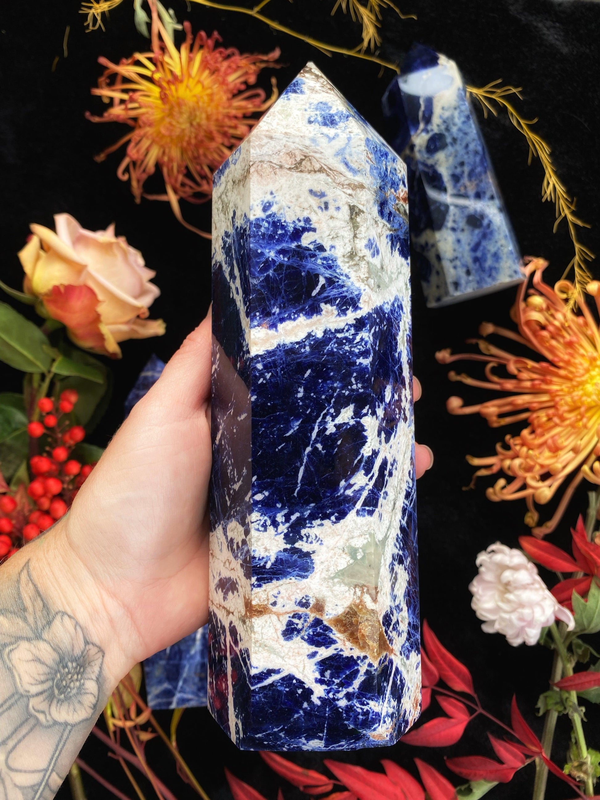 Blue crystal tower held by a hand with tattoos, surrounded by flowers on a dark background