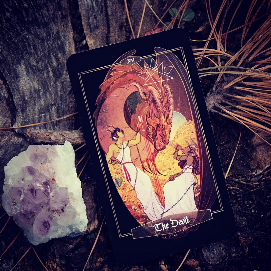 A tarot card deck featuring 'The Devil' card with a black background and an illustration of a red dragon and two figures in white robes, styled on a forest floor with pine needles, a crystal cluster, and a tree trunk.