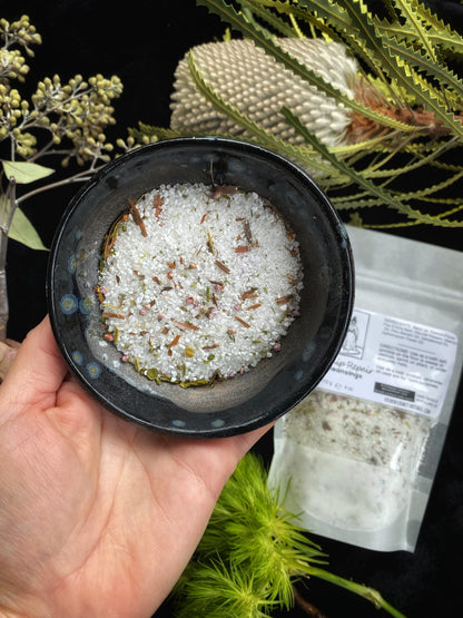Hand holding a small black container with white substance and small particles, surrounded by greenery.