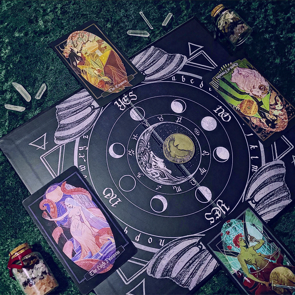 A tarot card deck and matching ritual mat laid out on a dark green textured surface. The central black circular mat features white moon phases, zodiac symbols, and a central moon illustration, surrounded by four illustrated tarot cards and small glass jars.