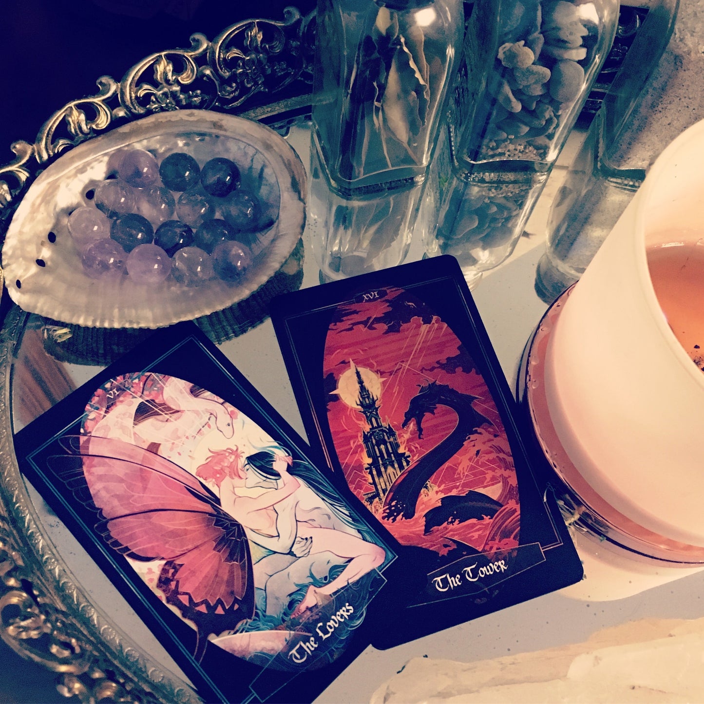 Two illustrated tarot cards, titled 'The Lover' and 'The Tower', displayed on a reflective surface next to a white candle, a shell of purple and blue stones, and glass containers.