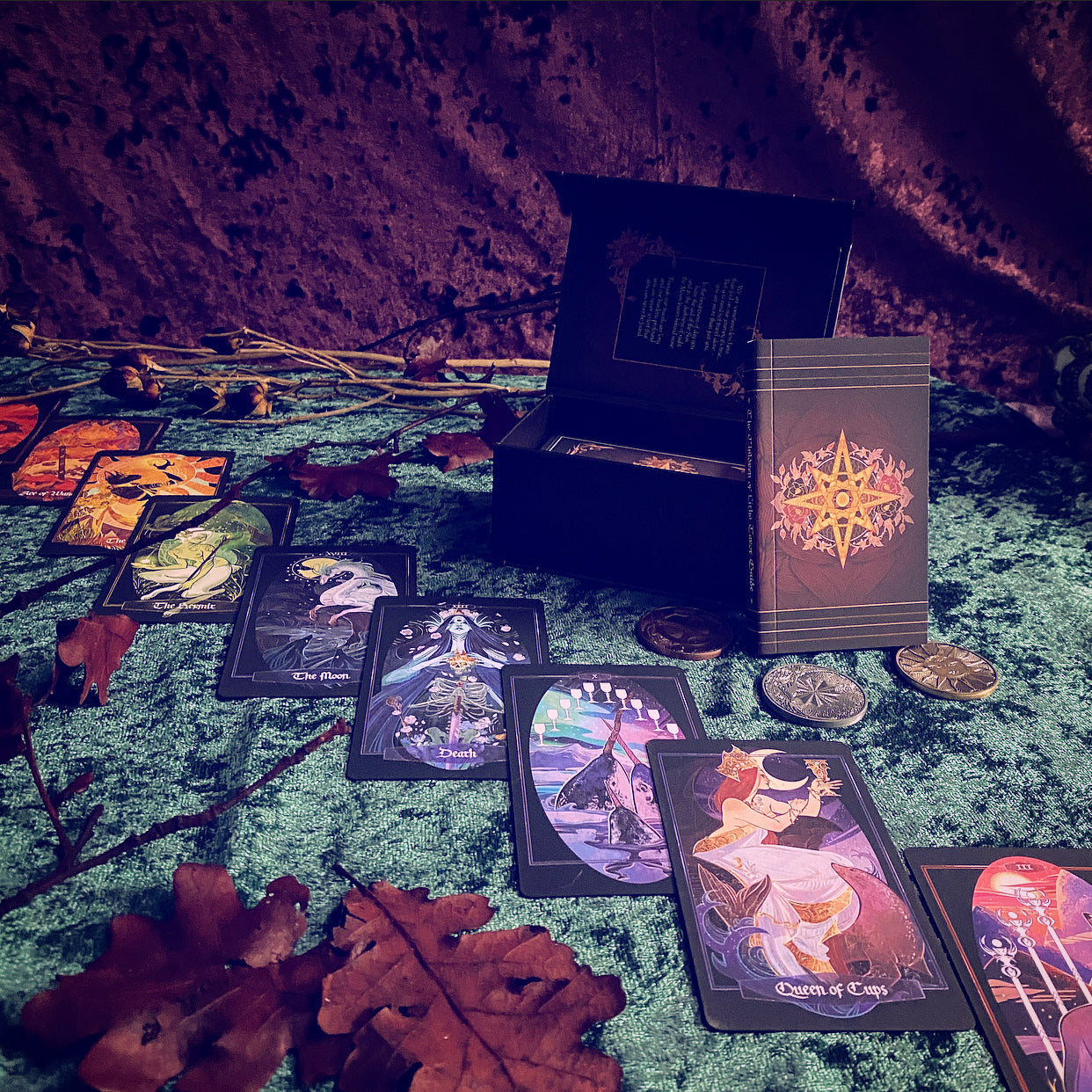 A tarot card deck displayed with an open black box, a standing card box, and several cards spread out on a mossy surface with autumn leaves. The cards feature colorful fantasy and mystical illustrations, and the scene is lit with a moody purple and orange glow.