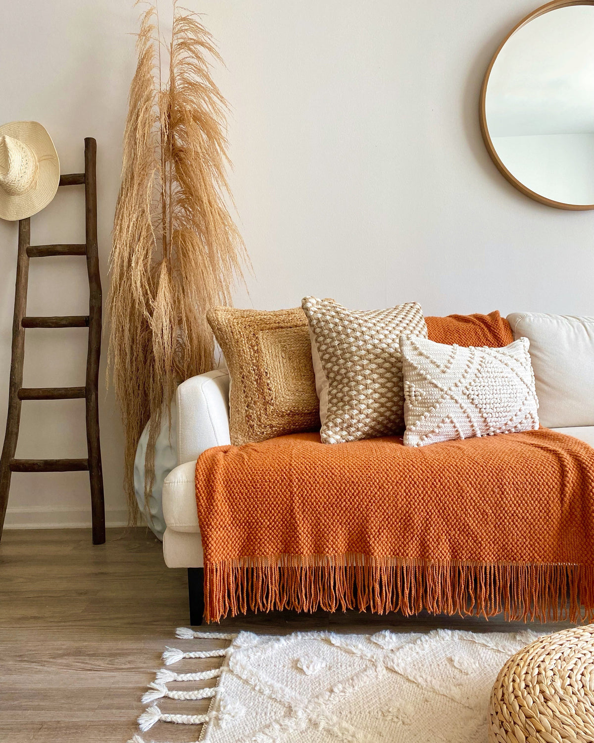 A cozy living room scene featuring a white sofa styled with a textured orange throw blanket, three neutral-toned decorative pillows, a tall dried pampas grass plant, a wooden ladder, and a round mirror on a light wall.