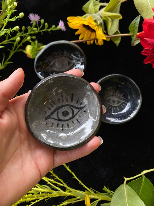 Hand holding a black ceramic dish with eye design against a dark background with flowers.