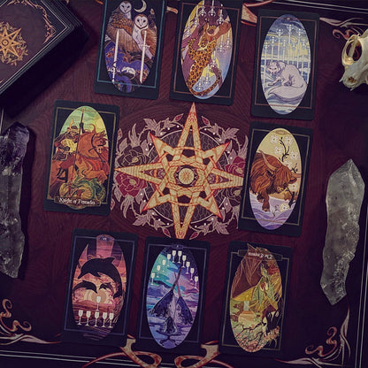 A styled flat-lay of tarot cards arranged on a dark red fabric background, featuring a large central pentagram with an eye motif. Surrounding cards show various illustrated scenes, and crystals and a decorative box are placed around them.