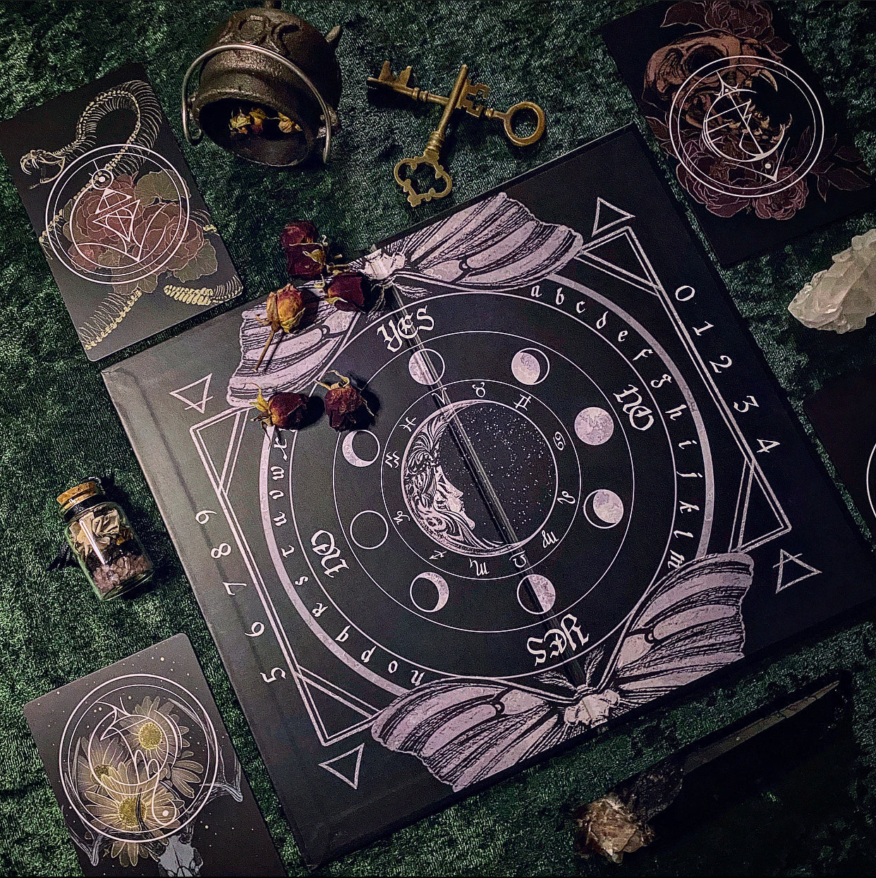 A dark occult-themed board game or ritual card set arranged on a textured green surface. The main square black board features moon phases, astrological symbols, and the words YES/NO and NORTHEAST/SOUTHEAST/etc.