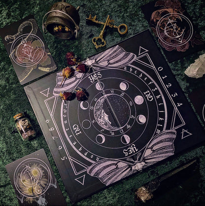 A dark occult-themed board game or ritual card set arranged on a textured green surface. The main square black board features moon phases, astrological symbols, and the words YES/NO and NORTHEAST/SOUTHEAST/etc.
