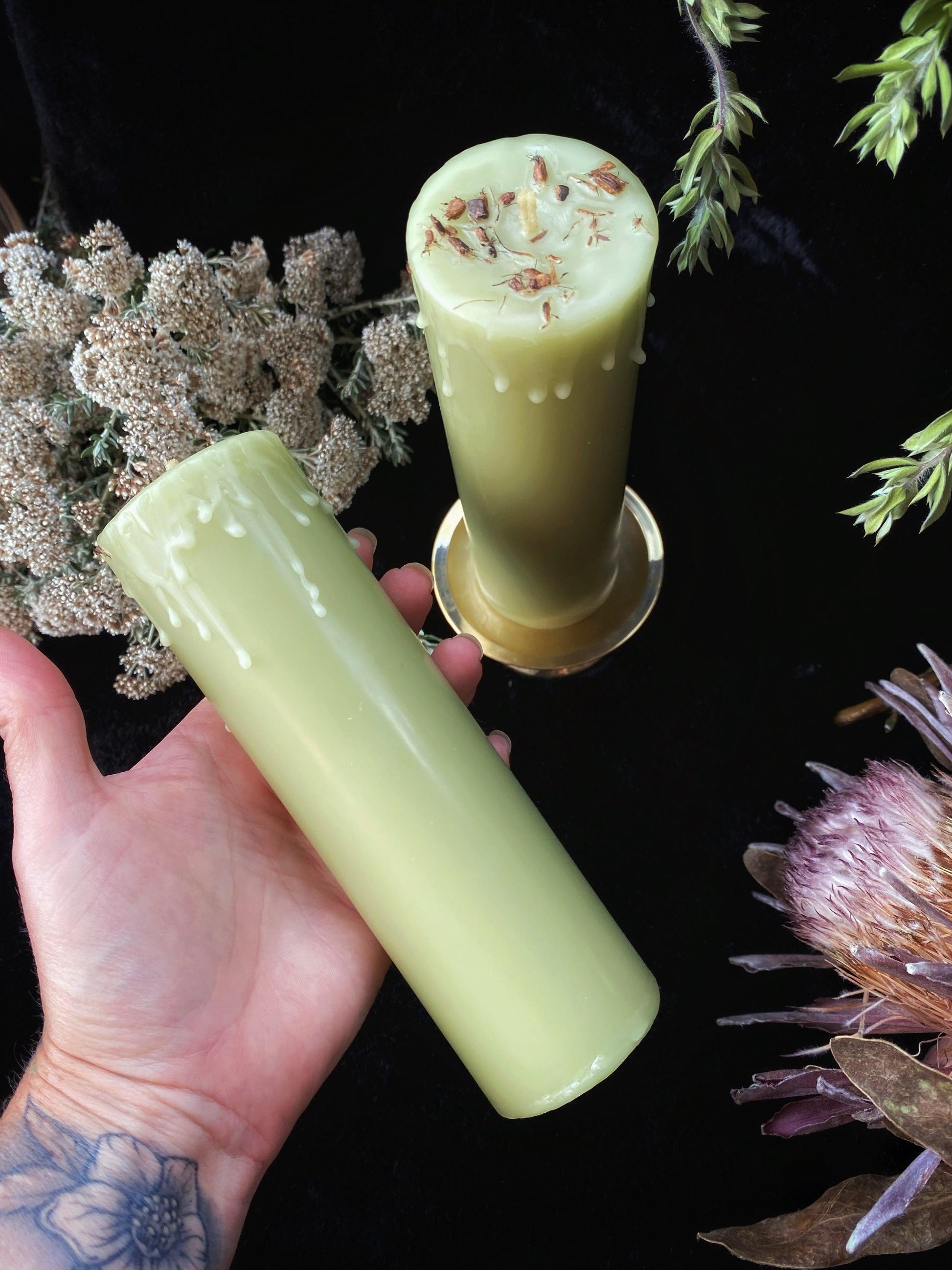 Two green candles, one held by a hand with tattoos, on a dark background with dried flowers.