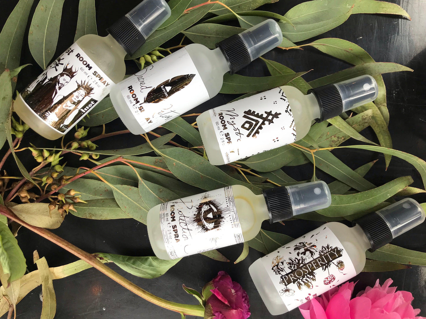 Multiple small bottles with labels on a dark surface with green leaves and pink flowers.