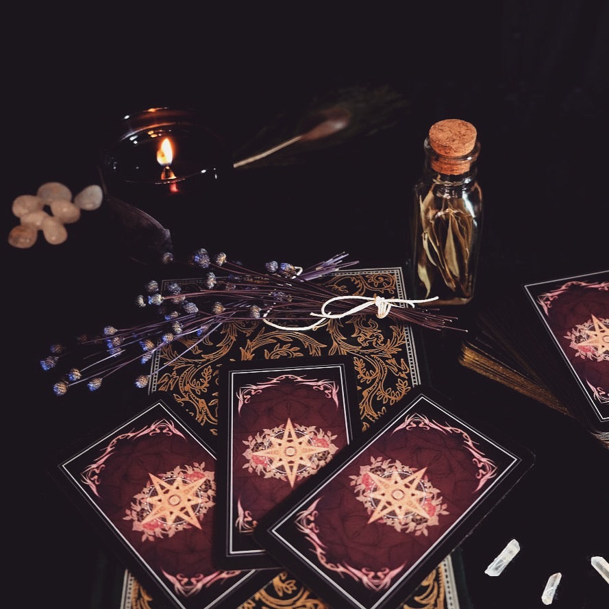 A styled flat-lay of tarot cards with a deep burgundy and gold design, arranged with dried lavender, a small glass bottle, crystals, and a candle on a dark background.