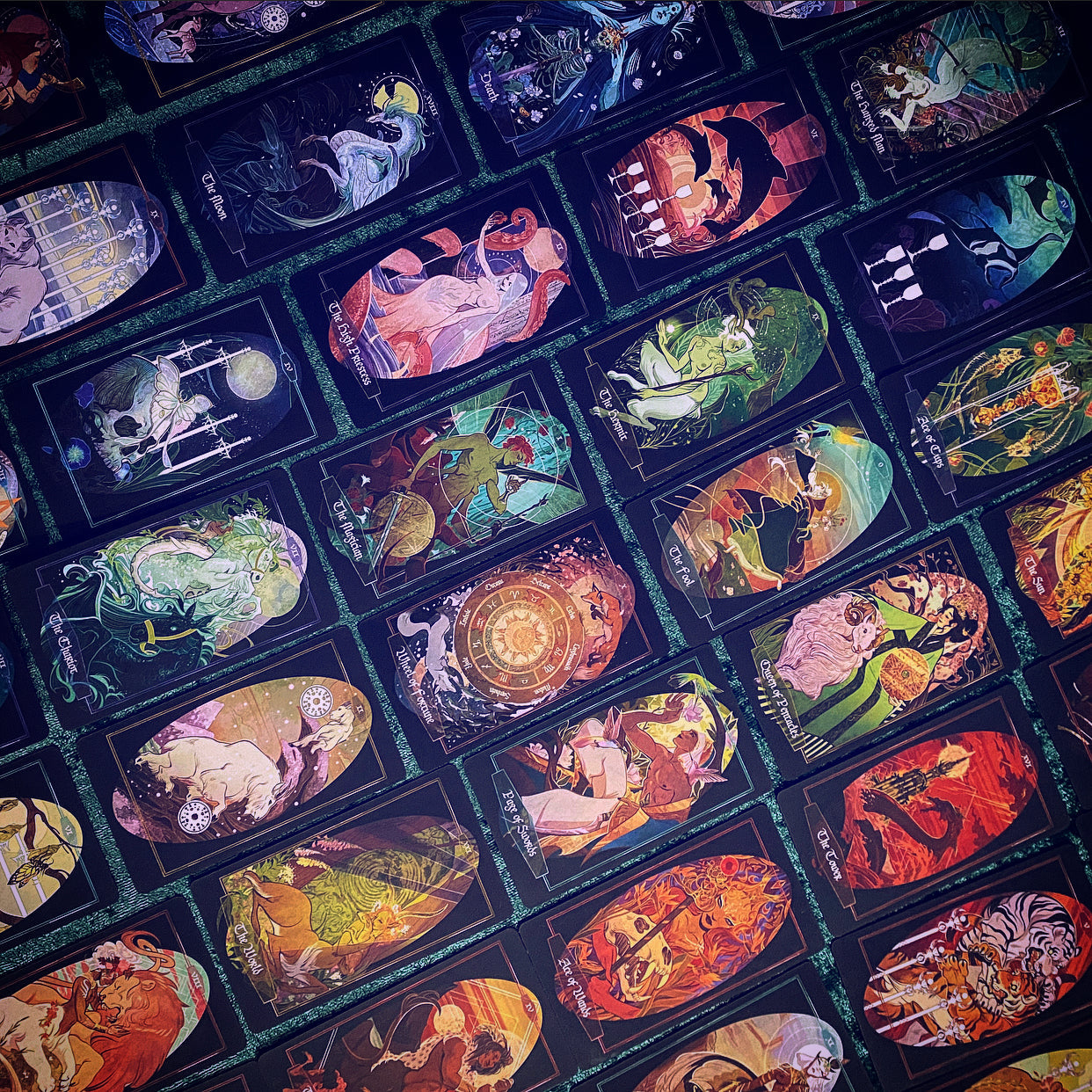 A close-up, angled view of a deck of illustrated tarot cards arranged in a grid. The cards feature highly detailed, colorful fantasy and mythological artwork on dark backgrounds, with teal borders separating the cards.