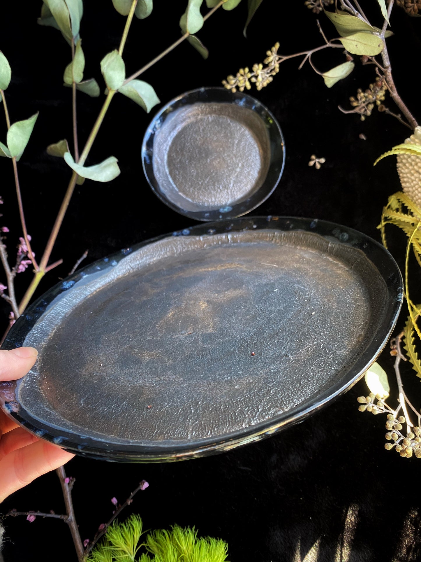 Black ceramic plates held against a dark background with greenery