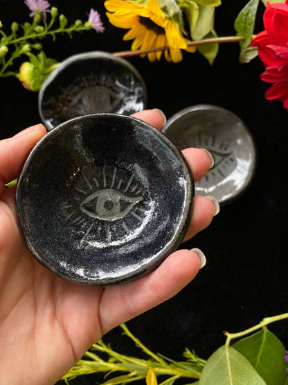 Hand holding a black ceramic dish with an eye design, surrounded by flowers on a dark background