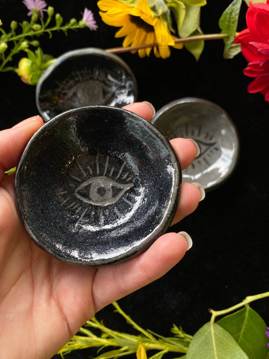 Hand holding a black ceramic dish with an eye design, surrounded by flowers on a dark background