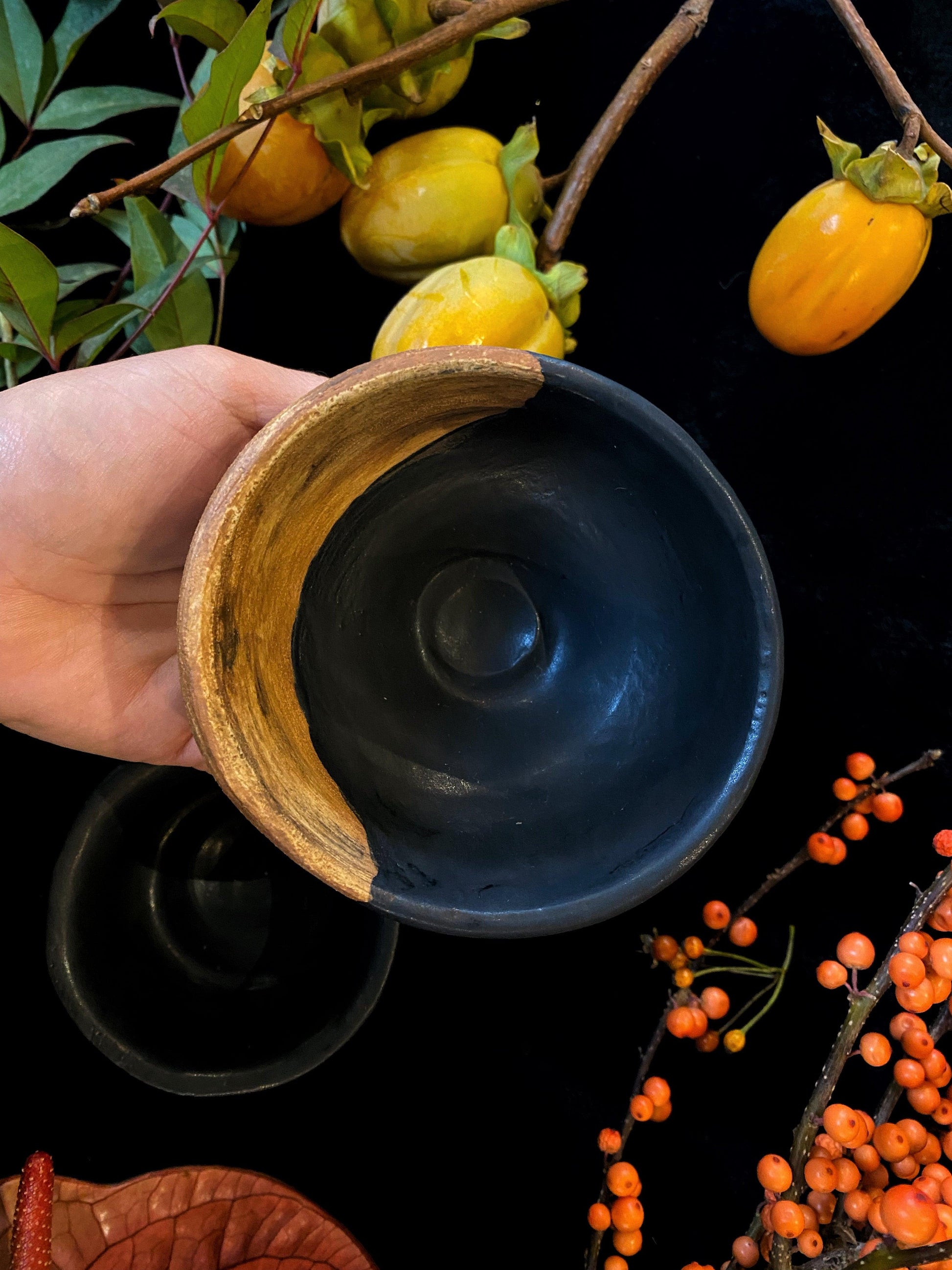 Hand holding a black ceramic bowl with wooden rim against a dark background with fruits and leaves.