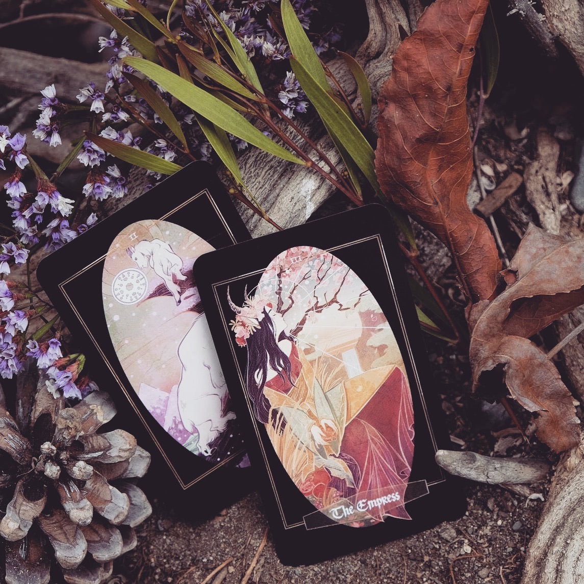 Two illustrated tarot cards, one labeled 'The Empress', arranged on a forest floor with dried leaves, pinecones, and small purple flowers.