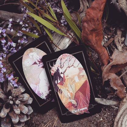 Two illustrated tarot cards, one labeled 'The Empress', arranged on a forest floor with dried leaves, pinecones, and small purple flowers.