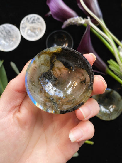 Hand holding a polished crystal with a blurred background of flowers and stones.