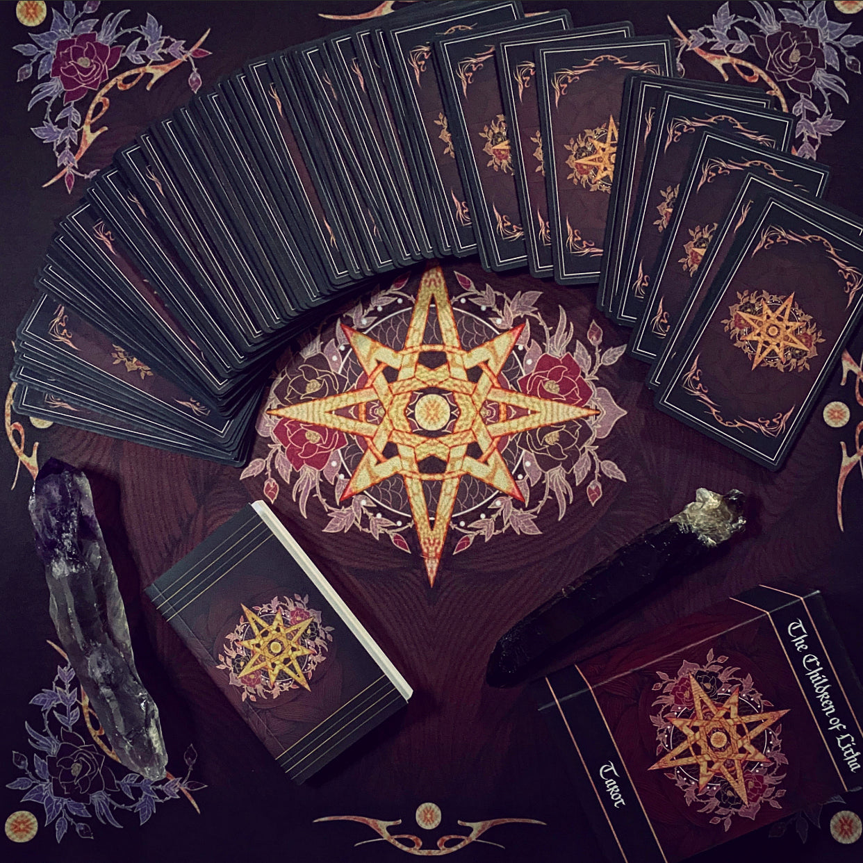 A styled flat-lay of a tarot deck with multiple cards fanned out, a closed deck box, a crystal ball, and a black wand on a dark patterned surface. The cards and box feature a prominent gold pentagram design with red floral accents on a dark background.