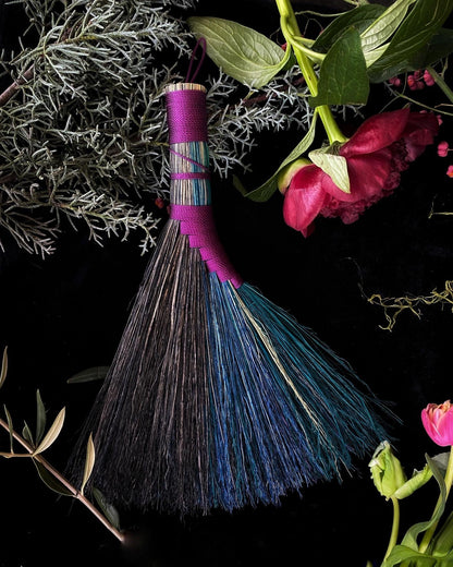 Broom with pink handle and blue bristles on a dark background with greenery