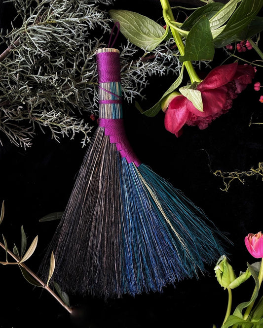 Broom with pink handle and blue bristles on a dark background with greenery