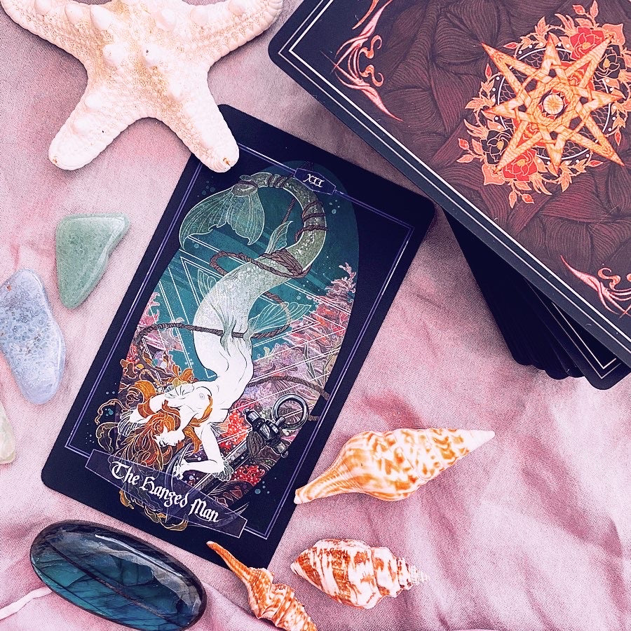 A tarot card deck styled in a flat lay on pink fabric with seashells, stones, and a star pendant. The main card shown is 'The Sargosed Man' with an underwater mermaid illustration, and another card deck is partially visible in the background.
