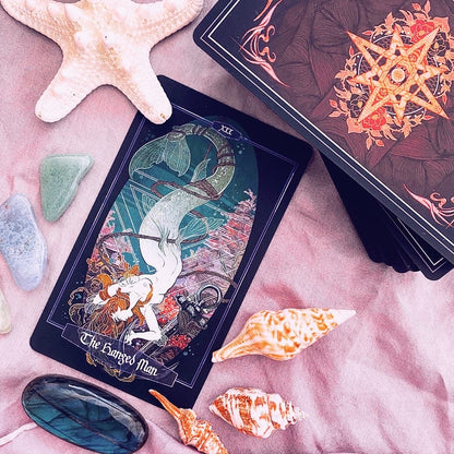 A tarot card deck styled in a flat lay on pink fabric with seashells, stones, and a star pendant. The main card shown is 'The Sargosed Man' with an underwater mermaid illustration, and another card deck is partially visible in the background.