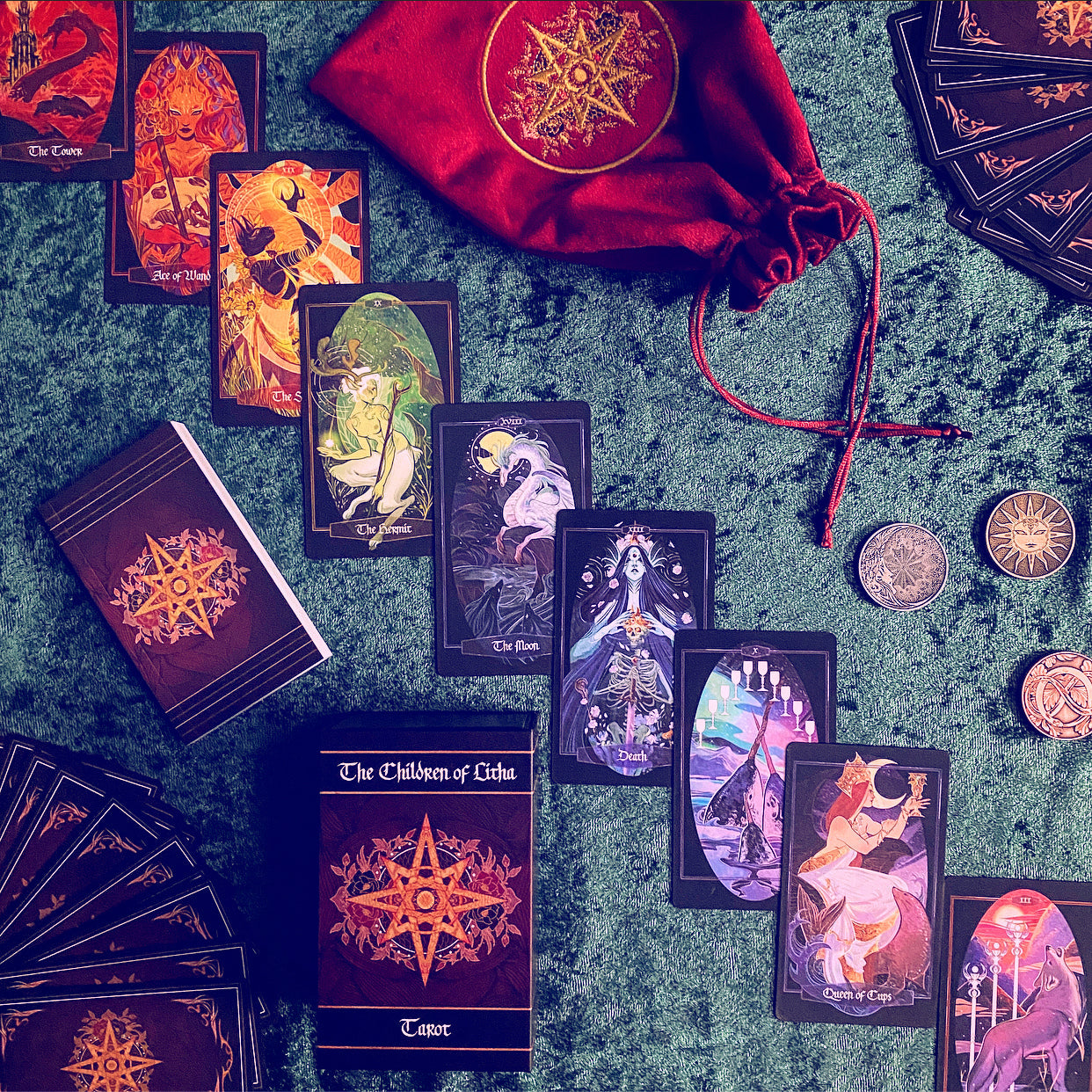A tarot card deck set arranged on a textured green surface, including multiple illustrated cards, a deck of cards, a red drawstring pouch with a gold emblem, and several metallic coins. The cards and pouch feature dark backgrounds with prominent gold star-like symbols and colorful fantasy-style artwork.