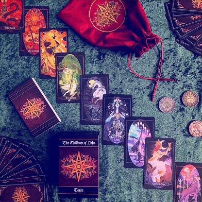 A tarot card deck set arranged on a textured green surface, including multiple illustrated cards, a deck of cards, a red drawstring pouch with a gold emblem, and several metallic coins. The cards and pouch feature dark backgrounds with prominent gold star-like symbols and colorful fantasy-style artwork.