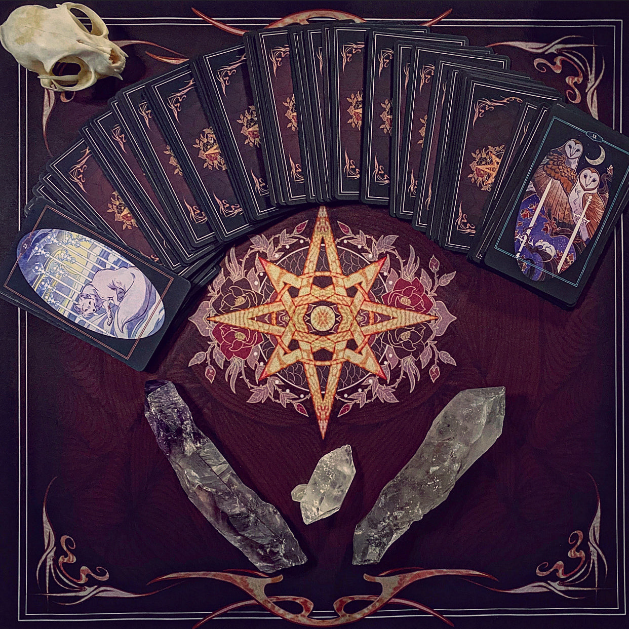 A tarot card deck arranged in a fan and stacked on a dark decorative mat with a central pentagram design, alongside a skull, crystals, and illustrated cards showing skeletal figures.