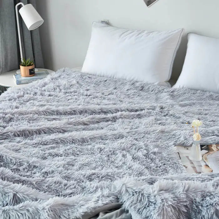 Gray fluffy blanket on a bed with white pillows and a lamp in the background.