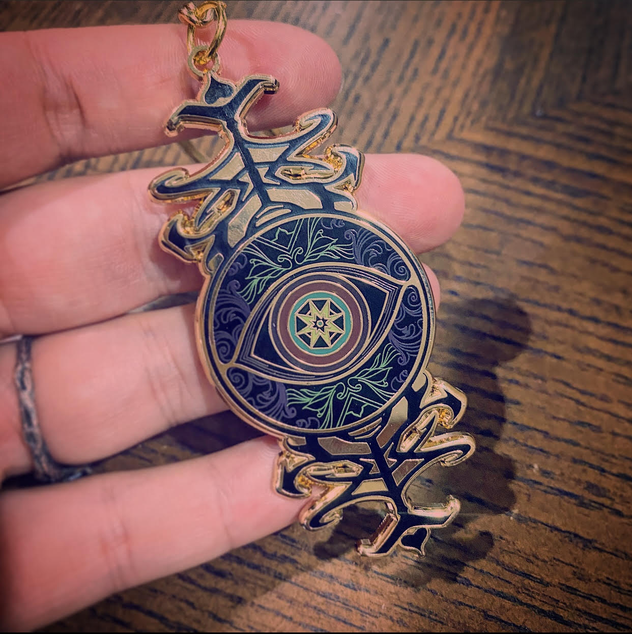 A hand holding a detailed circular pendant featuring an eye motif with gold, black, and blue enamel, set against a wooden tabletop background.