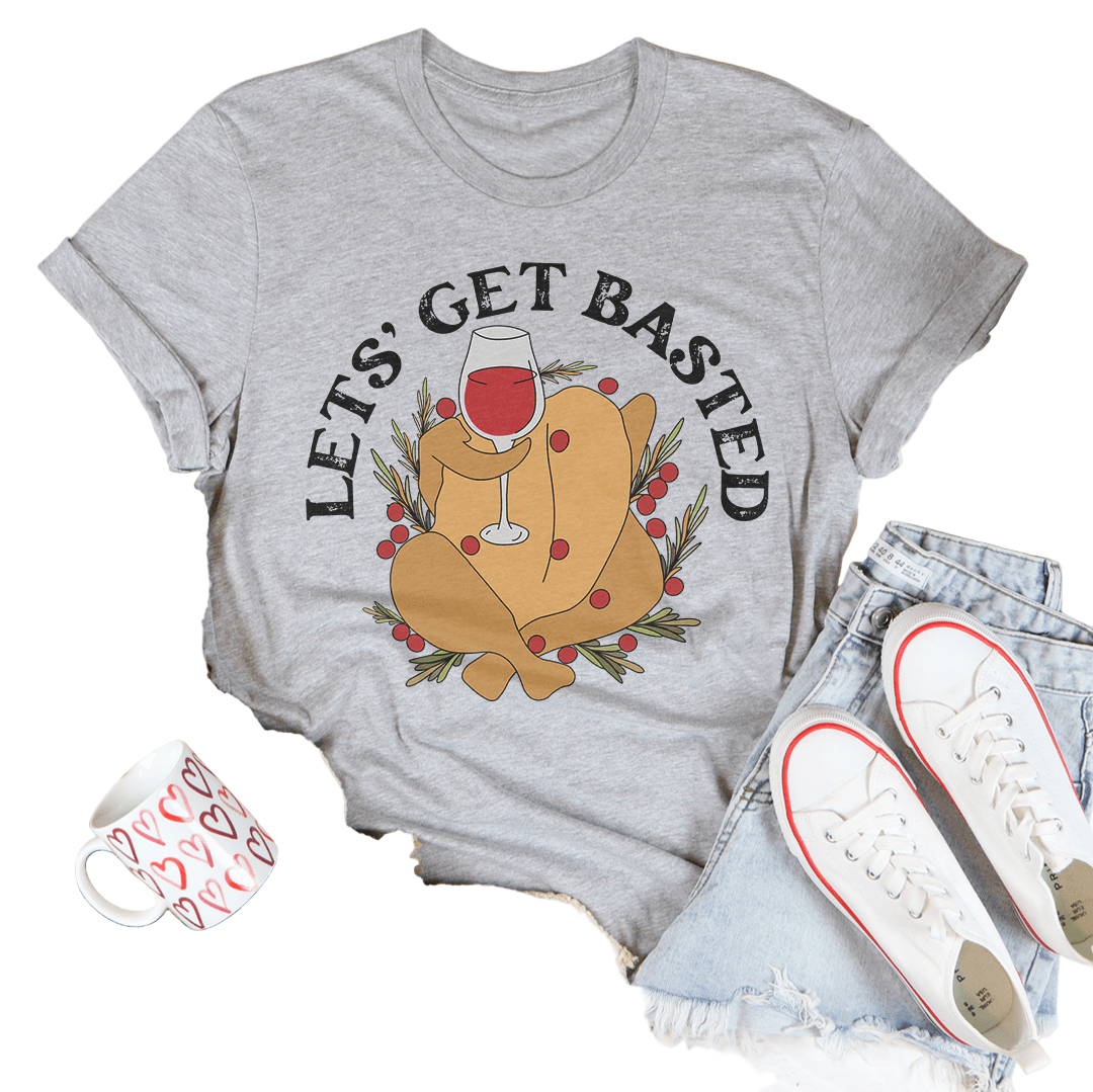 Gray t-shirt with a graphic of a roasted turkey and 'Let's Get Basted' text, surrounded by casual items on a light wooden surface.