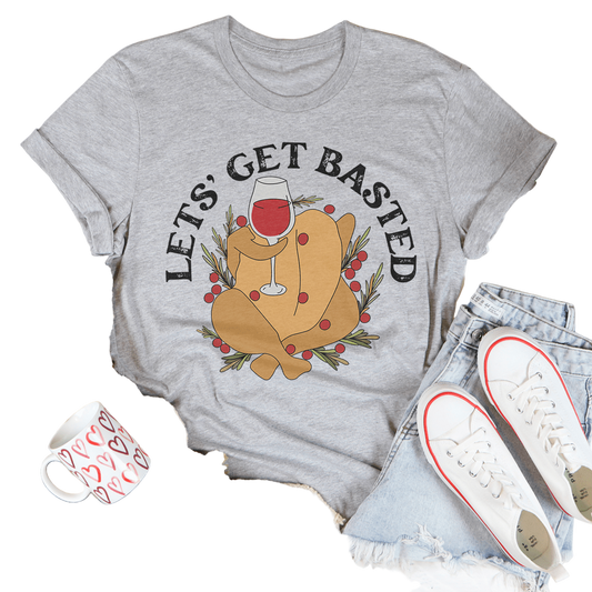 Gray t-shirt with a graphic of a roasted turkey and 'Let's Get Basted' text, surrounded by casual items on a light wooden surface.