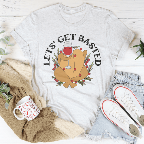 White t-shirt with a graphic of a roasted turkey and 'Let's Get Basted' text, surrounded by a mug, scarf, and sneakers on a light wooden surface.