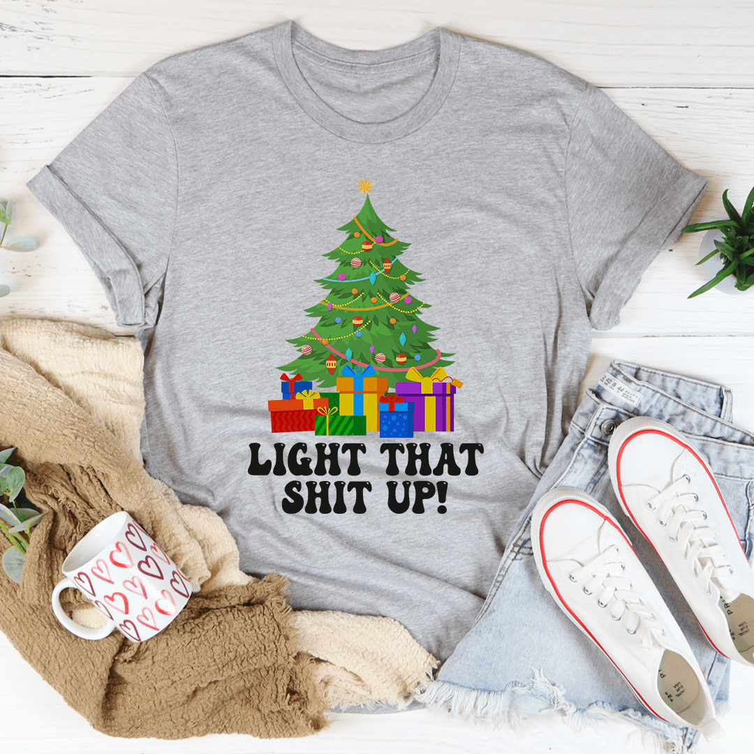 Gray t-shirt with Christmas tree and presents design, text 'LIGHT THAT SHIT UP!', surrounded by casual items on a light wooden surface.