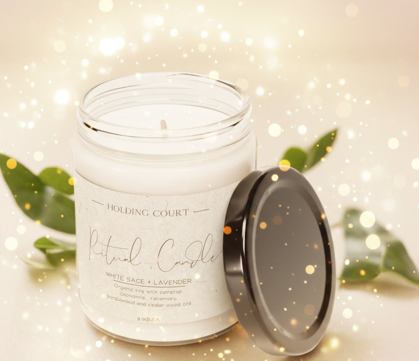 White candle in a glass jar with a black lid on a light surface, surrounded by green leaves.