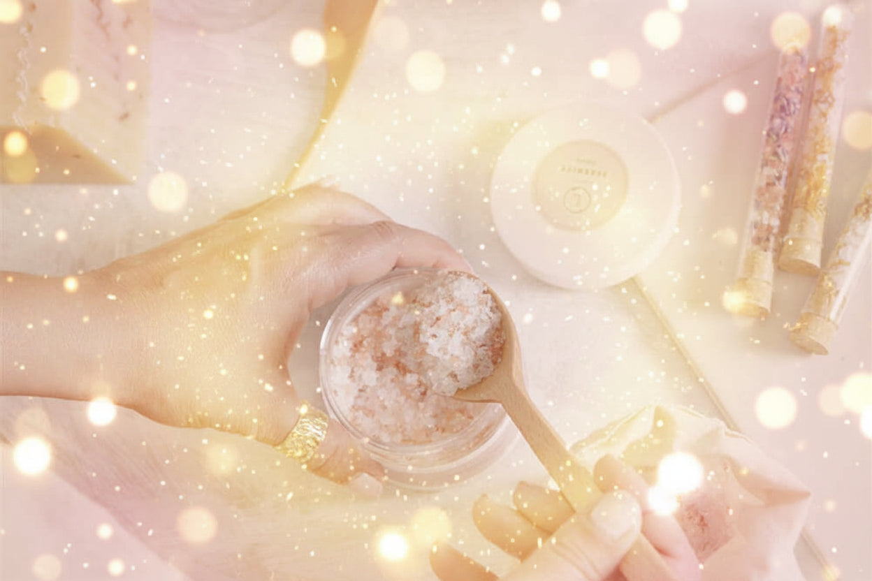Hand holding a wooden spoon with bath salt over a jar, surrounded by bath products on a light surface.
