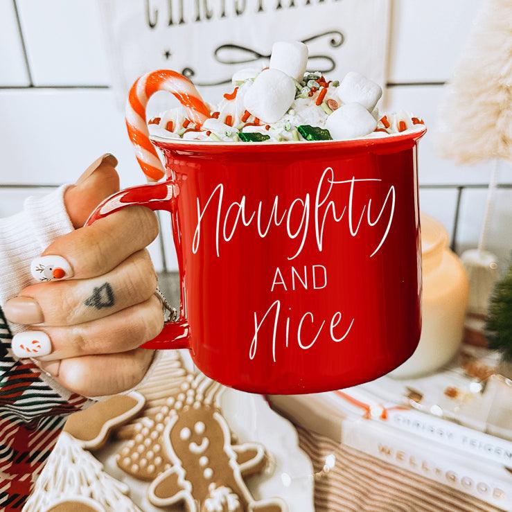 Red mug with 'Naughty and Nice' text, filled with hot chocolate and marshmallows, held by a person.