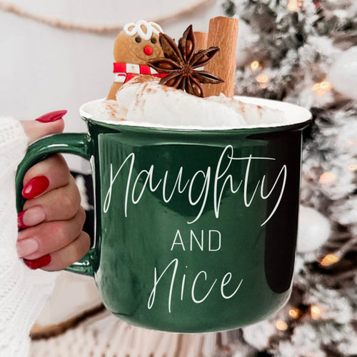 Green mug with 'Naughty and Nice' text, filled with hot chocolate and Christmas decorations, held by a person.