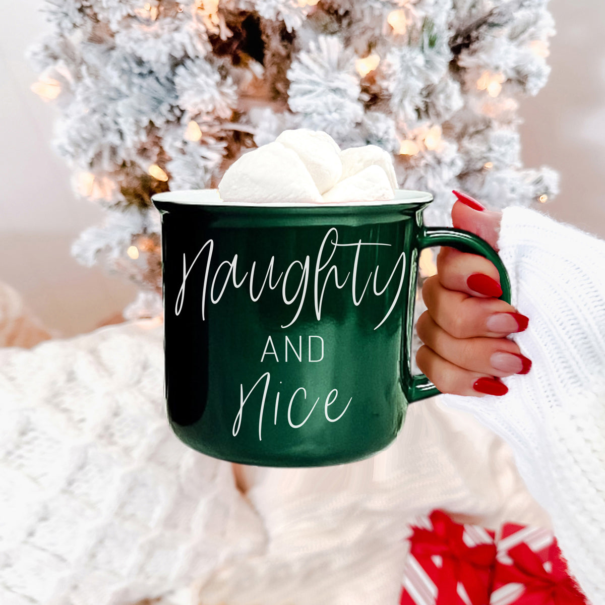 Green mug with 'Naughty and Nice' text held by a person in front of a decorated Christmas tree.
