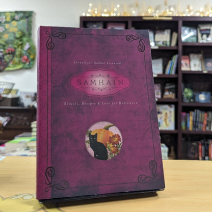 A hardcover book titled "SAMHAIN" from the "Aromantic & Lesbian Essentials" series, featuring a textured purple cover with a central oval illustration of a black cat and pumpkins, displayed on a wooden table in a library setting.