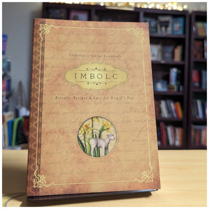 Book titled 'Imbolc' with decorative cover featuring a goat and flowers, set against a bookshelf background.