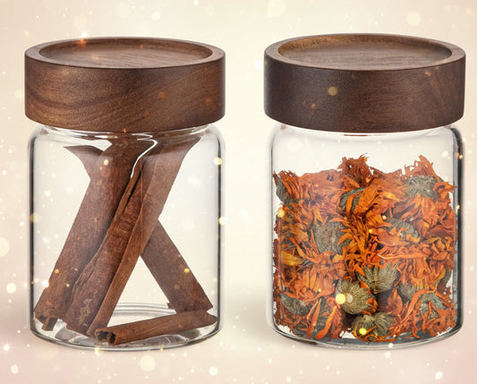 Two glass jars with wooden lids, one containing cinnamon sticks and the other dried herbs.