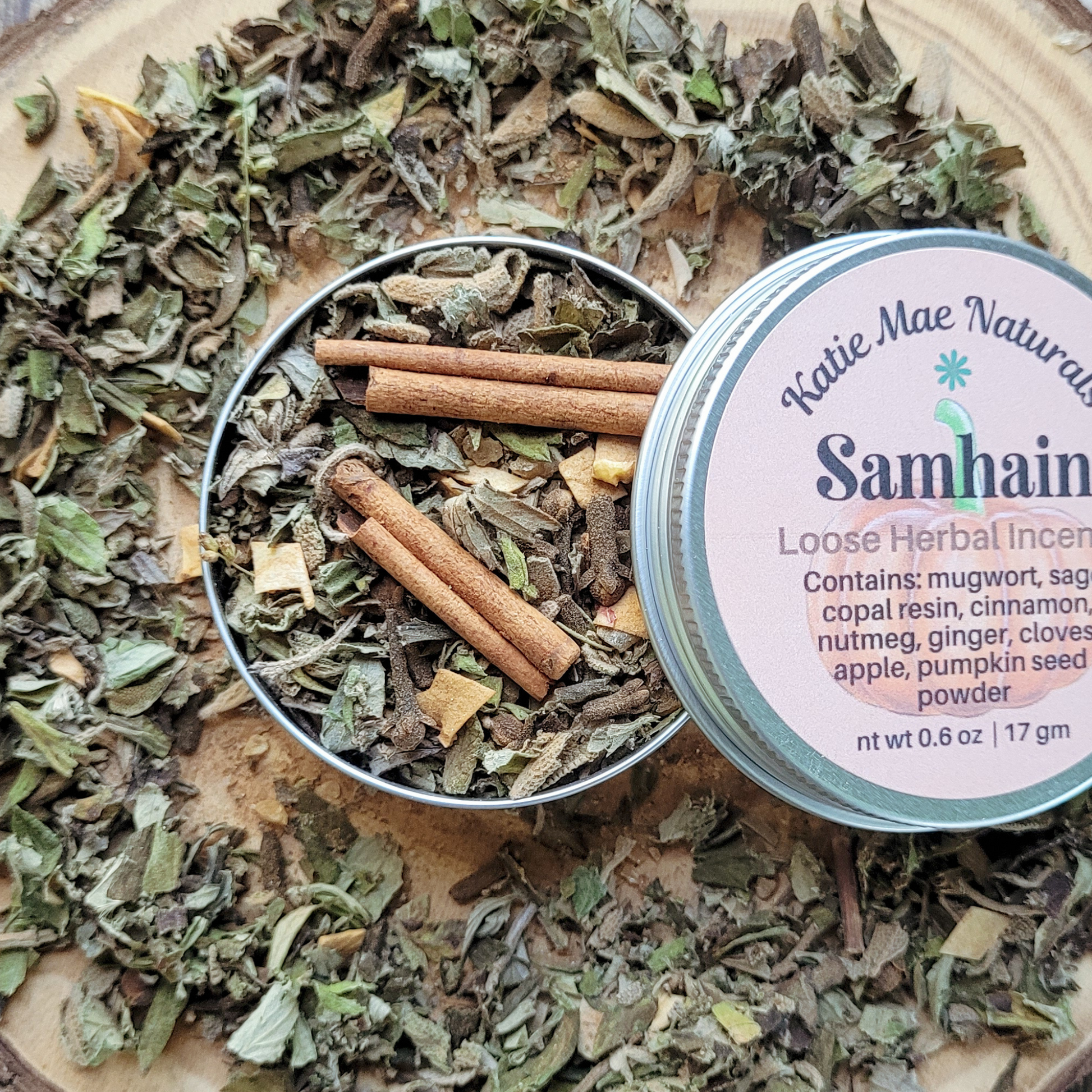 Loose herbal incense with cinnamon sticks and a container labeled 'Katie Mae Naturals Samhain' on a wooden surface.