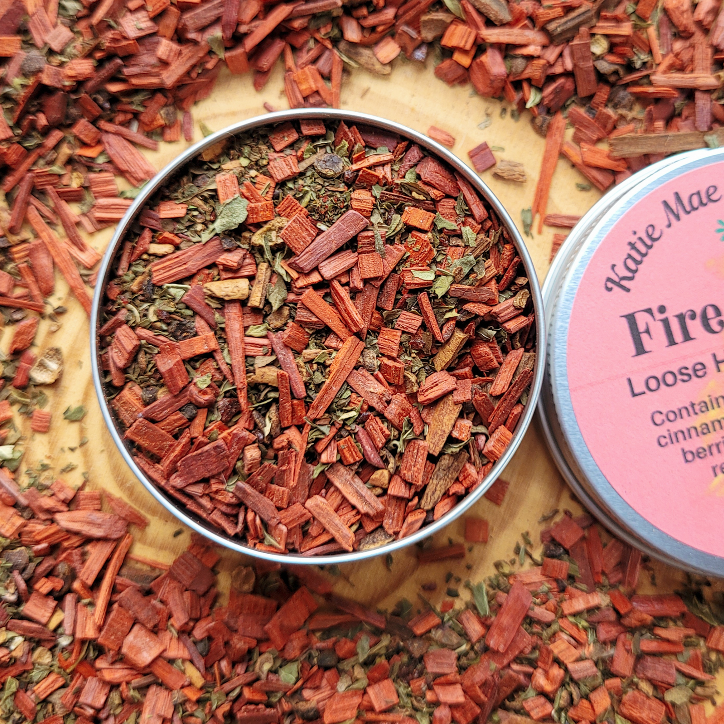 Metal tin filled with red and brown dried herbs on a bed of similar herbs, with a pink label visible.