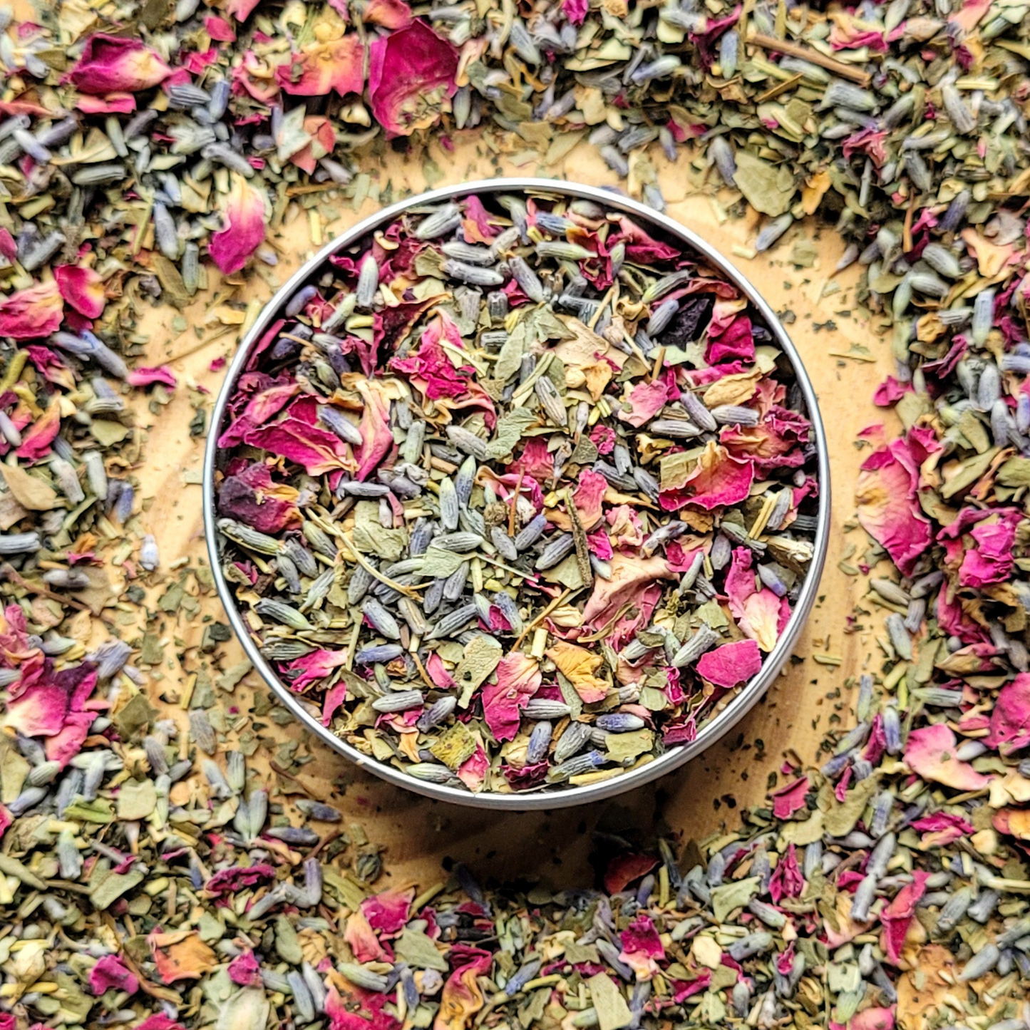Colorful dried herbs and flowers in a metal container surrounded by more herbs on a wooden surface
