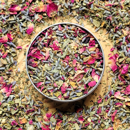 Colorful dried herbs and flowers in a metal container surrounded by more herbs on a wooden surface